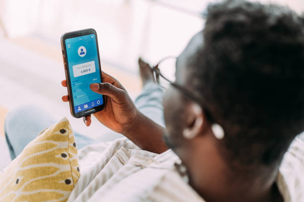 Young man using mobile app on smart phone for checking his finances while sitting on the sofa at home. Shot of young man using smartphone for checking his money balance of the Internet online wallet while relaxing on sofa in living room. man looking at phone over shoulder stock pictures, royalty-free photos & images