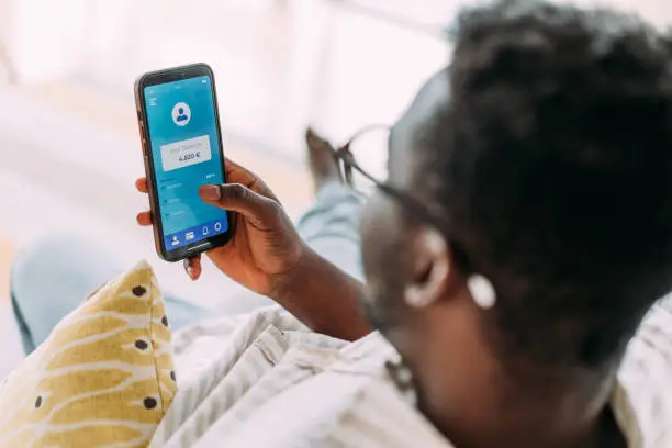 Photo of Young man using mobile app on smart phone for checking his finances while sitting on the sofa at home.