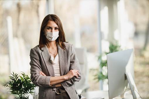 A successful young business woman with surgical mask standing in her office and looking at camera.