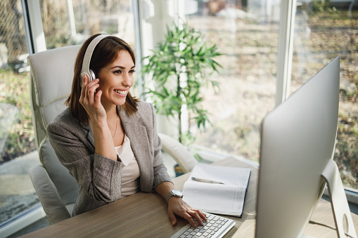 A smiling business woman sitting alone in her home office with headphones and working online on computer.