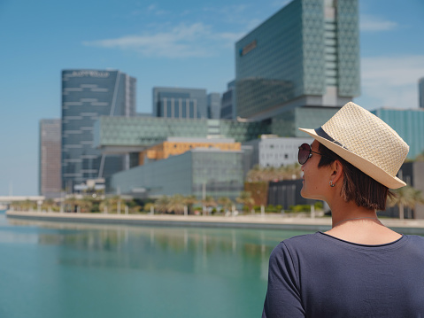 travel to the United Arab Emirates, Happy young asian female traveler in dress and hat look at View of Abu Dhabi Skyline in Al Maryah Island. Vacation and tourist destination concept.