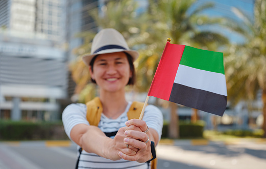 Happy young asian female traveler with backpack and hat with UAE flag against scenic skyscrapers in Abu Dhabi. Immigration, student education and arab emirates citizenship concept