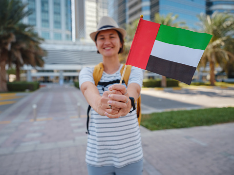 Happy young asian female traveler with backpack and hat with UAE flag against scenic skyscrapers in Abu Dhabi. Immigration, student education and arab emirates citizenship concept