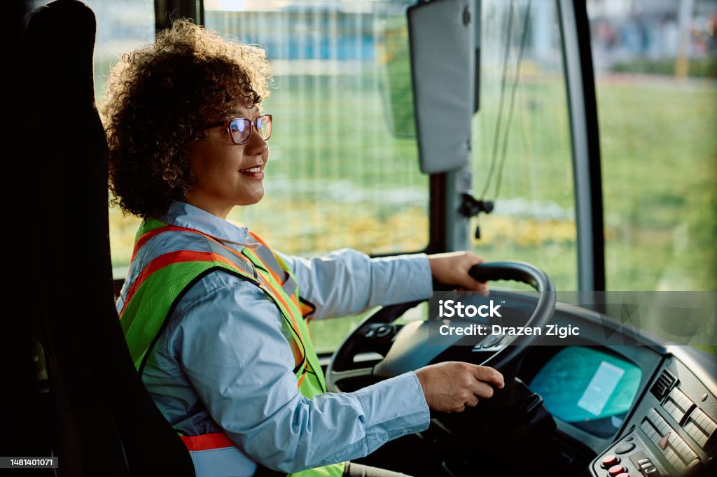Happy woman driving a bus in reverse while looking at rear view mirror. Happy female bus driver looking at rear view mirror while parking in reverse at the station. Bus Driver Stock Photo