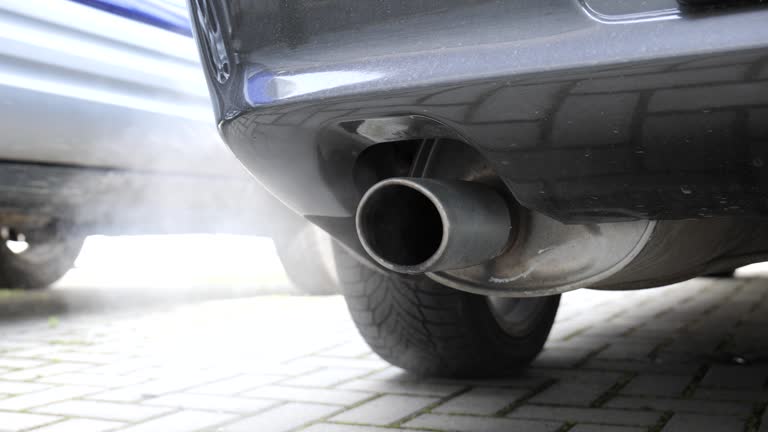 Car tailpipe or exhaust pipe tip emitting smoke. Standing nondescript car, low angle close up view, real time, no people