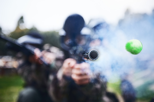 Paintball, shooting and person with a gun during a game, competition or match on a field. Fire, smoke and tool in motion for attack, battle and aim with a weapon during an outdoor cardio sport