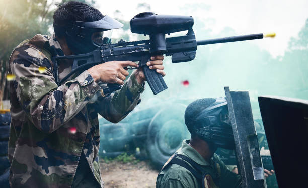 training, paintball and men with shield and gun in camouflage safety gear at military game target practice. teamwork, shooting sports and war games, play with rifle and working together at army park. - paintballing violence exercising sport imagens e fotografias de stock