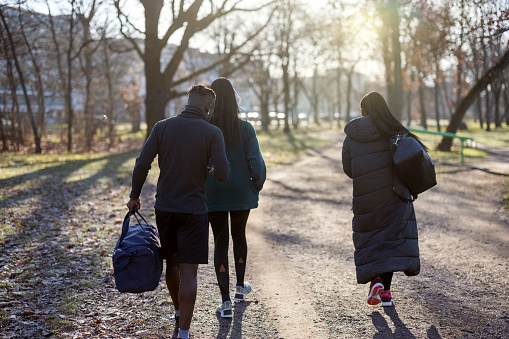 Rear view of three young people walking to gym with duffle bags. Fitness people walking through city park with gym bags on an autumn morning.
