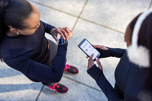 Overhead view of two young women standing together using their smart devices after a workout session outdoors. African female friends checking their fitness progress on smartphone and smartwatch after training session.
