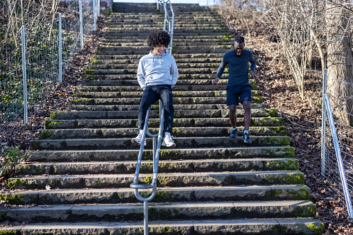 Two young male friends having fun while exercising at a park. African man running down the steps with a friend sliding down a railing at the park.