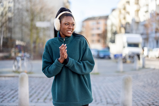 Portrait of African woman in sportswear and ear muff standing by city street after workout. Young female feeling cold after morning run outdoors in the city.