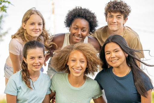 A small group of teens huddle together outside as they pose for a portrait.  They are each dressed casually and are smiling.