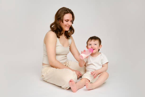 Happy baby with mother drinks milk from bottle on studio white background. Resting child with mom eats formula. Kid about two years old (one year nine months)