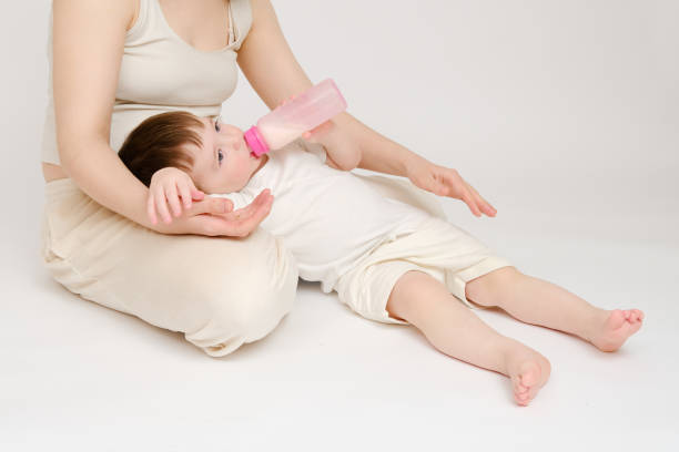 Happy baby with mother drinks milk from bottle on studio white background. Resting child with mom eats formula. Kid about two years old (one year nine months)