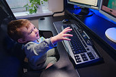 Happy baby is sitting at the computer keyboard with a bottle of milk in his hands. A child with a desktop PC in a home living room with milk formula from a bottle. Kid aged one year nine months