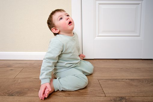 Little baby coughs sitting on the cold laminate floor in the house. Small child is sick and coughing in the home living room. Kid aged one year nine months