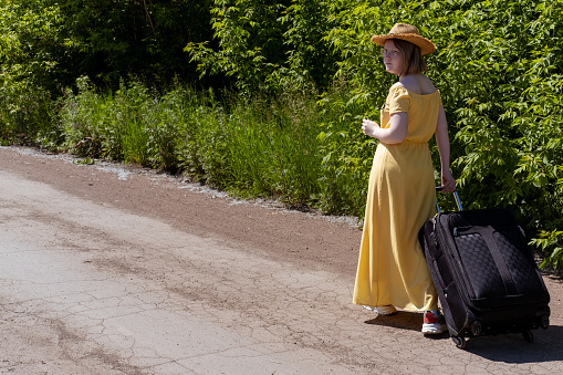 Asian teenage girl in a yellow dress, glasses and a hat walking along a rural dirt road with a suitcase. Ecotourism. Summer time. Holidays. Travel concept