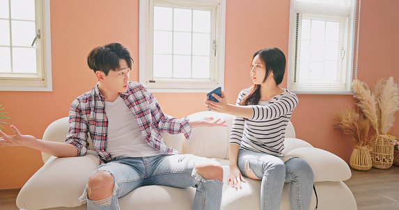 asian young couple argument concept - asian woman holding mobile phone is asking man about some mistrust or distrust and jealousy problems when they sitting on couch at home