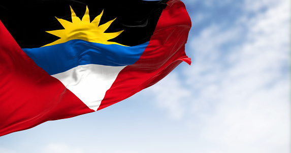Antigua and Barbuda national flag waving on a clear day. Red field with inverted triangle of black, blue, white and rising sun. 3d illustration render. Fluttering textile