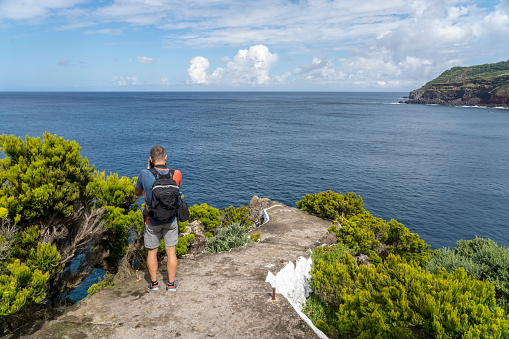 Rear men standing on the edge of the coast of island Terceira photographing the view at the blue sea on sunny day. Azores.