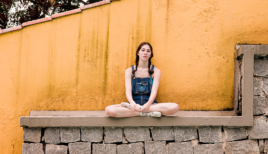 Teenager girl sitting alone on a rocky wall with yellow background.