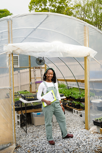 A farmer stands in front of her greenhouse and smiles at the camera.  She is wearing green overalls and a long sleeved white shirt.  She is black, has shoulder length dreadlocks tied back with a a green headband, and is holding a clipboard.  You can see various seedlings growing in the greenhouse behind her.