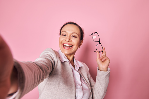 Selfie of a multi-ethnic attractive woman holding stylish trendy eyeglasses, smiling with a beautiful toothy smile looking at camera on isolated pink background. Pretty brunette making self-portrait