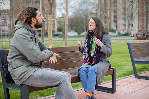 Two young deaf people are hanging out in the public park. They are sitting on the park bench and speaking in sign language.