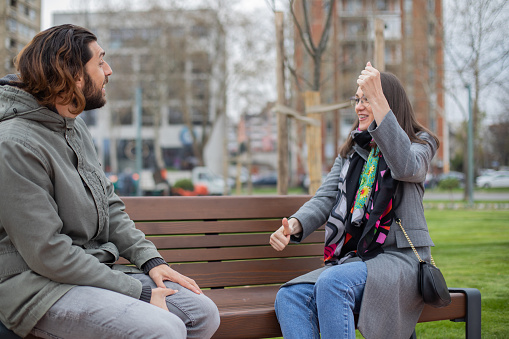 Two young deaf people are hanging out in the public park. They are sitting on the park bench and speaking in sign language.