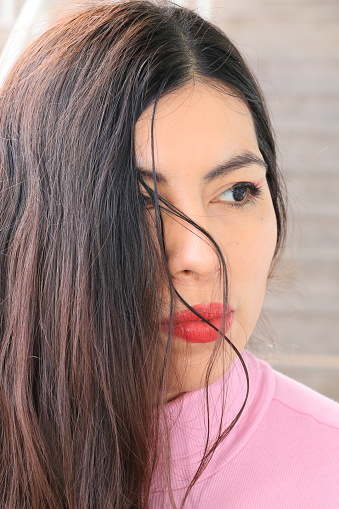 A closeup of a Mexican woman. She is wearing makeup, long brown straight hair over one eye, and a pink sweater.