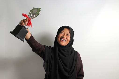 Clever young Asian girl student in black hijab or headscarf raising her trophy as an achievement from winning in school competition isolated on white background