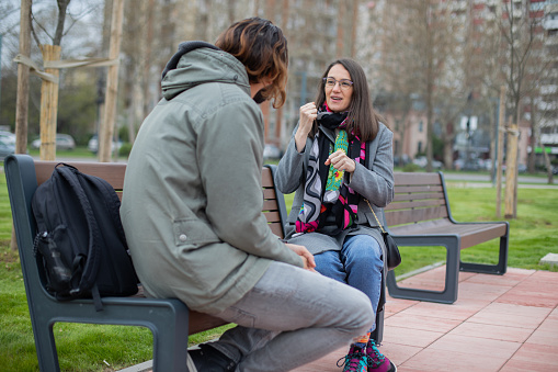 Two young deaf people are hanging out in the public park. They are sitting on the park bench and communicating in sign language.