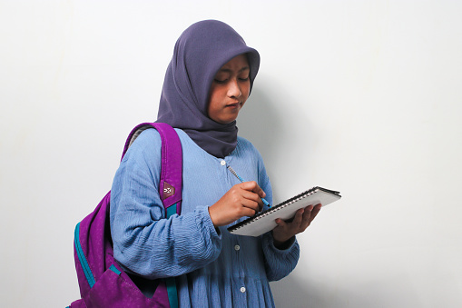 Diligent young Asian girl student wearing hijab and backpack, writing something on the book isolated on white background.