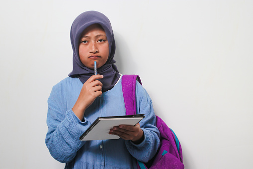 Thoughtful young Asian girl student in hijab wearing backpack interested in thinking while holding a book and pen isolated on white background