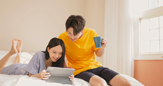 asian young couple is looking some information together on social media in the internet through digital tablet at bedroom home happily - man drinking beverage with mug while woman lying bed