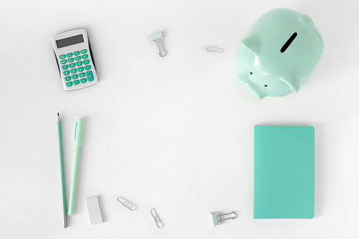 Top view of white desk with stationery. Office accessories in blue green colors. Creative concept with piggy bank. Horizontal flat lay of supplies and copy space.