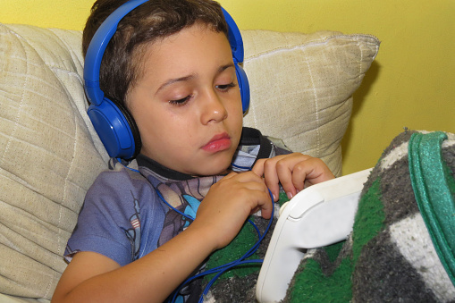 Boy lying down with headphones having fun playing games on his graphic tablet. The photo was taken inside a house.