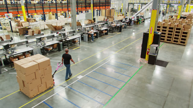 Aerial View Of  Man Pulling Dolly Full Of Boxes Past Packing Stations In Fulfillment Center
