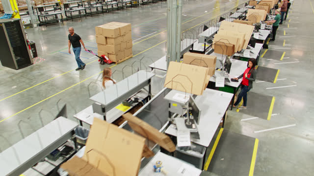 Aerial Shot of Man Pulling Dolly Full Of Boxes In Fulfillment Center Past Packing Stations