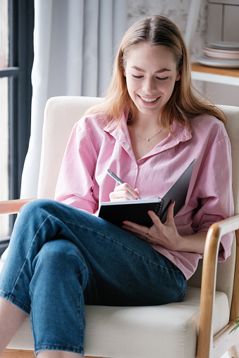 Laughing caucasian young woman in casual clothes is sitting at home in a chair with her legs crossed, making notes in a planer, writing a story in a book