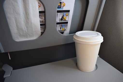 Plain white disposable coffee cup on a grey foldable airplane tray table.