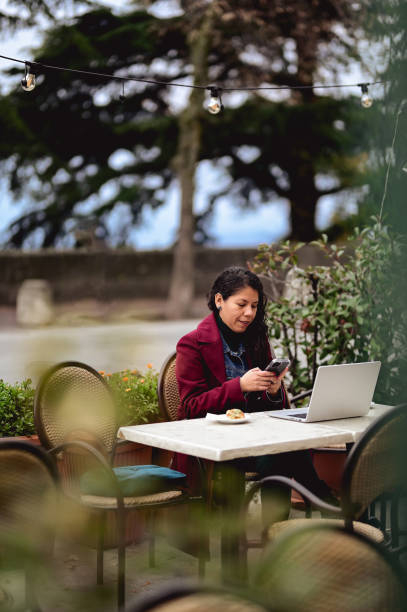 istock latin mid adult female scrolling her smart phone while sitting on the table and working on laptop. 1481459544