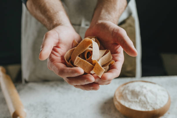 istock Unrecognizable man in an apron holding dough cut into strips on a white table against a dark wall 1481466219