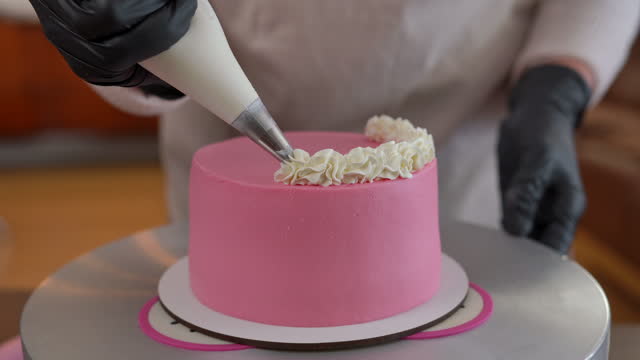 Close-up unrecognizable female baker turning pink baked cake decorating with white butter cream topping. Young woman baking delicious pastry indoors.