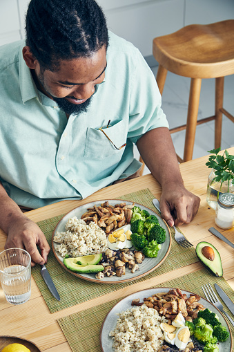 A man preparing a healthy protein lunch, after the workout, made of chicken meat, brown rice, mushrooms,Spanish onion, tomatoes, seeds and broccoli, served in a modern plate on a home wooden table top, representing a healthy lifestyle and wellbeing, an image with a copy space