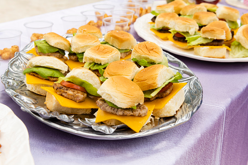 This is a close up photograph of platters of ready to eat cheeseburger sliders on a buffet table outdoors.