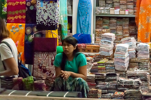 Mandalay, Myanmar - nov 3, 2012: a young woman sells colorful fabrics on a stall in the covered market in Mandalay.