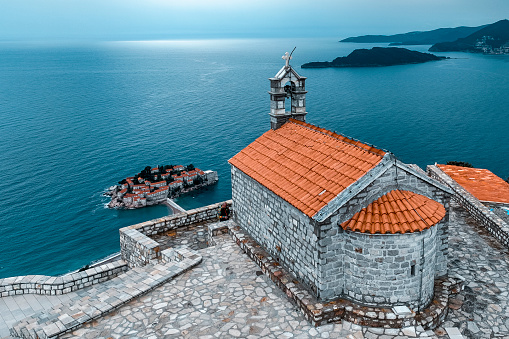 Old church of St. Sava in a small village of Blizikuce captured with a drone and with Sveti Stefan island in a background during one cloudy afternoon in the Adriatic sea.
