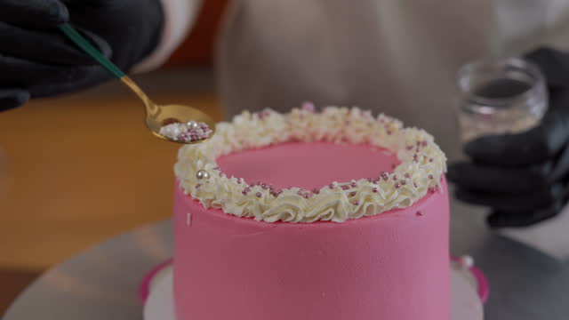 Close-up sprinkling edible pearls on round pink cake with white butter cream decoration. Unrecognizable woman decorating delicious pastry indoors close-up.