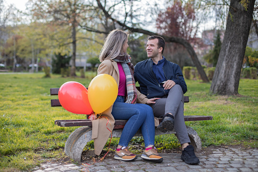 Beautiful young couple are enjoying in the public park. They are sitting on the park bench , talking and holding balloons.